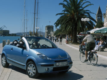 Een blauwe Citroën C3 Pluriel cabriolet staat geparkeerd op een zonnige boulevard aan het water, met een fietser en buitendiners in de buurt. Palmbomen en historische gebouwen bevinden zich op de achtergrond.