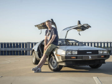 Een persoon leunt tegen een DeLorean met de iconische vleugeldeuren open, geparkeerd in een industriële omgeving onder een heldere hemel.