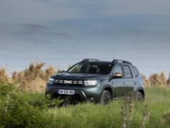 Een Dacia Duster Extreme met lopende koplampen rijdt op een bewolkte dag door een grasveld.