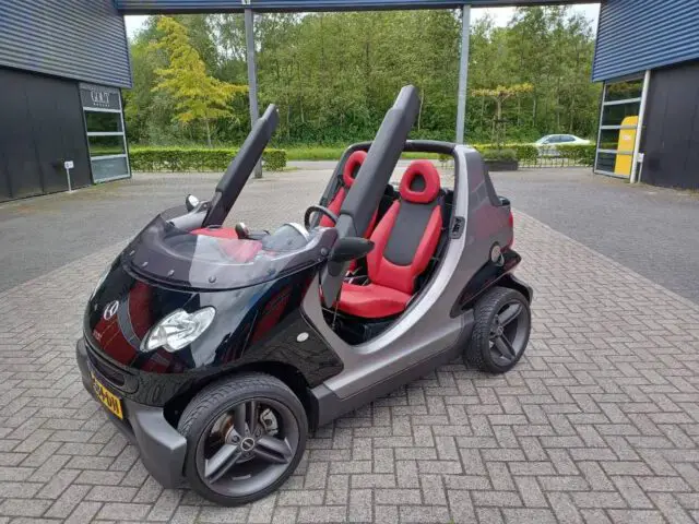 Een kleine tweezits cabrio met een zwart-zilveren carrosserie, rode stoelen en geen deuren staat geparkeerd op een verhard oppervlak met gebouwen en groen op de achtergrond.