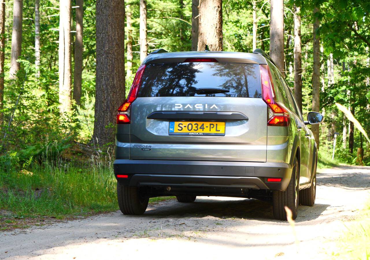 Een grijze Dacia Jogger Hybrid rijdt over een onverharde weg door een bosrijk gebied. Op de achterste kentekenplaat van het voertuig staat "S-034-PL".