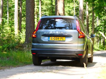 Een grijze Dacia Jogger Hybrid rijdt over een onverharde weg door een bosrijk gebied. Op de achterste kentekenplaat van het voertuig staat "S-034-PL".