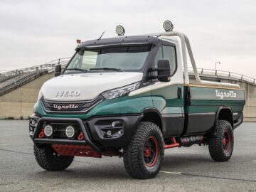 Een groen-witte IVECO Daily 4x4 Tigrotto-truck met offroad-banden en grilleverlichting staat geparkeerd op een asfaltoppervlak nabij een industrieel bouwwerk.
