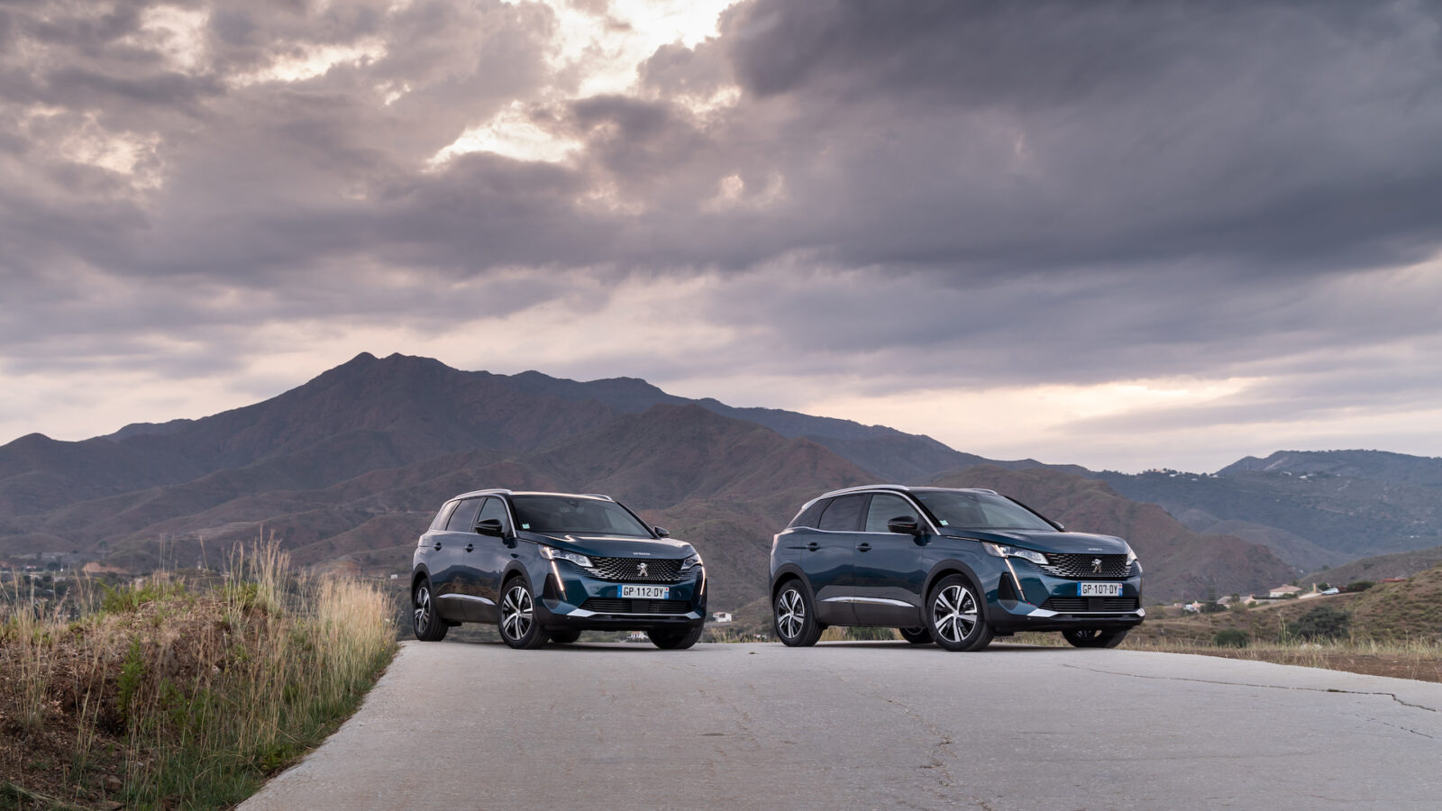 Twee identieke donkergekleurde Peugeot 5008 48V HYBRID SUV's staan geparkeerd op een betonnen weg met bergachtig terrein en een bewolkte lucht op de achtergrond.