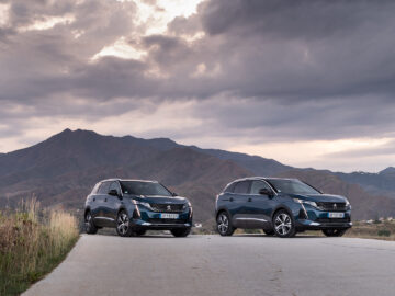 Twee identieke donkergekleurde Peugeot 5008 48V HYBRID SUV's staan geparkeerd op een betonnen weg met bergachtig terrein en een bewolkte lucht op de achtergrond.