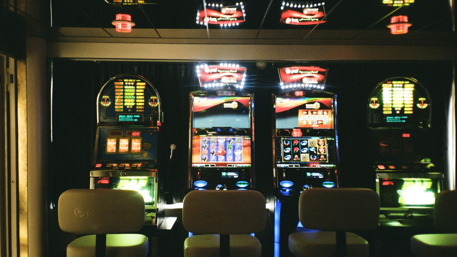 Een rij verlichte gokautomaten met verschillende displays en vier lege stoelen ervoor, geplaatst in een slecht verlicht casino, belicht enkele van de populairste casinospellen.