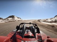 Een rode raceauto, bestuurd door Robin Shute, navigeert over een bochtige bergweg met een besneeuwd landschap en helderblauwe lucht op de achtergrond. De zon schijnt fel.