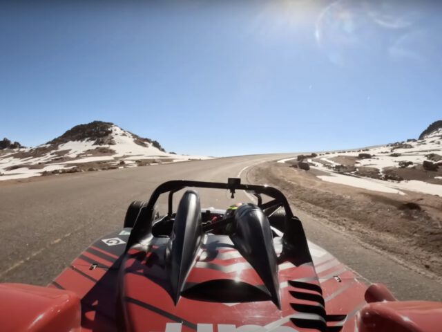 Een rode raceauto, bestuurd door Robin Shute, navigeert over een bochtige bergweg met een besneeuwd landschap en helderblauwe lucht op de achtergrond. De zon schijnt fel.