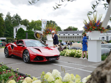 Een rode sportwagen die te zien is tijdens het Wheels at the Palace-evenement, omgeven door mensen, bloemstukken en groen, met een wit gebouw en spandoeken op de achtergrond.