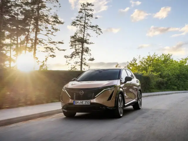 Een bronskleurige elektrische Nissan ARIYA SUV met Europees kenteken rijdt over een weg, omlijst door bomen en een ondergaande zon op de achtergrond. Dit pittoreske tafereel geeft de essentie weer van reizen in de leasewereld.