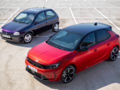 Op een leeg perceel staan twee hatchback-auto's geparkeerd. De auto op de voorgrond is een moderne rode Opel Corsa met een zwart dak, terwijl de auto op de achtergrond een ouder model is in donkerpaars.
