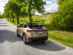 Een bronzen Nissan ARIYA SUV rijdt op een verharde, landelijke weg omzoomd door bomen en grasvelden onder een gedeeltelijk bewolkte hemel. Nissan verrassende leasewereld met deze schitterende elektrische auto, beschikbaar voor € 549,- per maand.