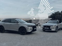 Twee moderne zilveren SUV's, versierd met het DS-logo, staan geparkeerd op een grindoppervlak met een schilderachtige achtergrond van heuvels en bomen. Een digitale afbeelding van witte geometrische lijnen is over het luchtgedeelte achter de voertuigen heen gelegd.