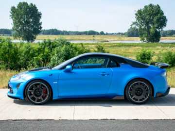 Een blauwe Alpine A110R staat geparkeerd op een verharde weg met een graslandschap en bomen op de achtergrond, wat de essentie van zijn sportieve elegantie weerspiegelt.