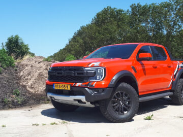 Een felrode Ford Ranger Raptor met zwarte accenten en grote terreinbanden staat geparkeerd op een betonnen ondergrond met groen en een zandheuvel op de achtergrond.