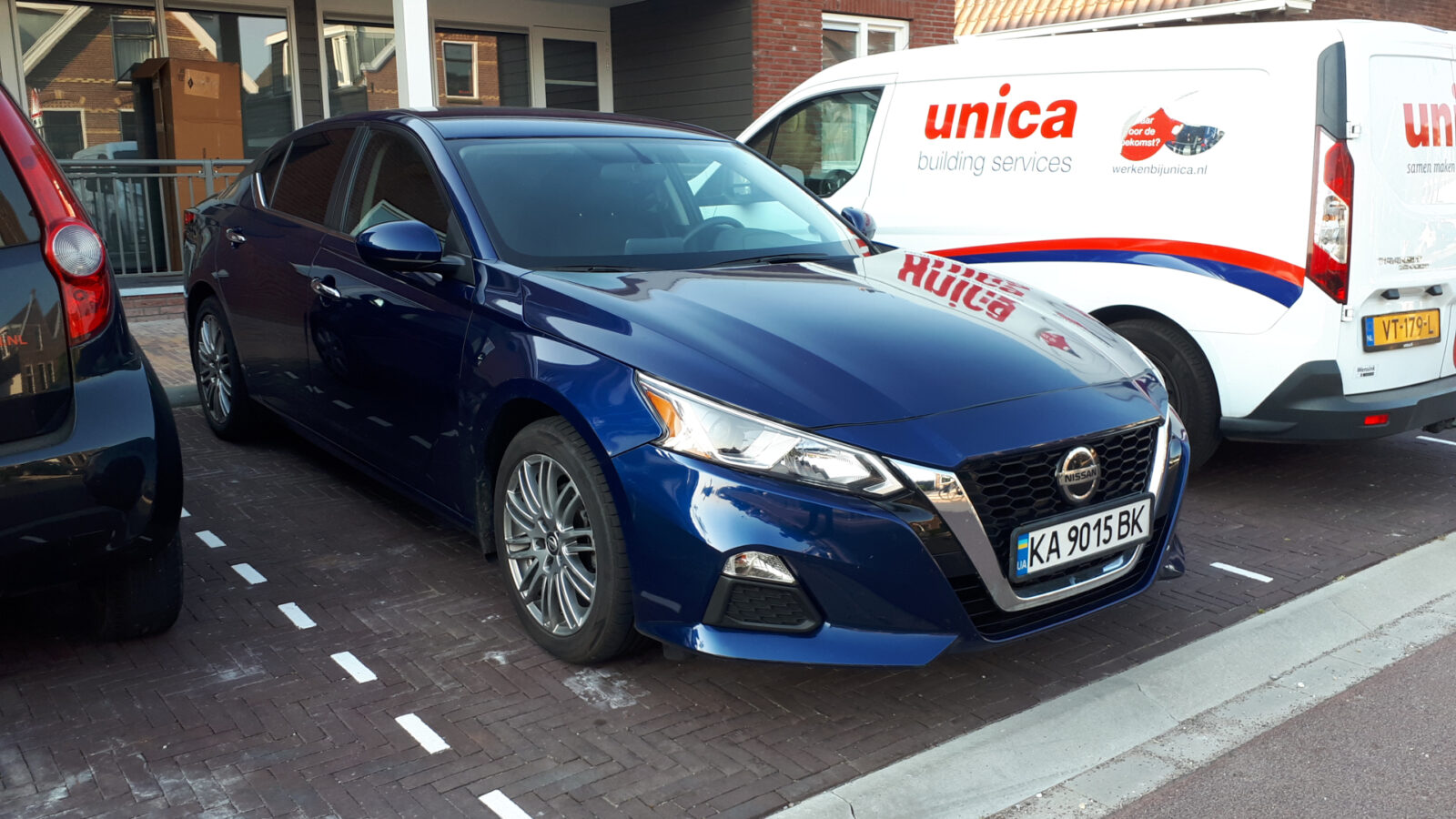 Op een gemarkeerde parkeerplaats staat een blauwe Nissan Altima geparkeerd naast een witte Unica-busje, waardoor een beeld ontstaat van het dagelijks leven buiten het gebouw.