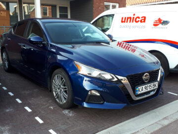 Op een gemarkeerde parkeerplaats staat een blauwe Nissan Altima geparkeerd naast een witte Unica-busje, waardoor een beeld ontstaat van het dagelijks leven buiten het gebouw.