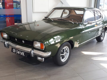 Een groene klassieke Ford Capri-coupé wordt binnenshuis tentoongesteld op een tegelvloer, met de linkervoorzijde naar de camera gericht. De auto, voorzien van een Nederlands kenteken met de tekst "01-34-VK", straalt de tijdloze charme van de Ford-techniek uit.