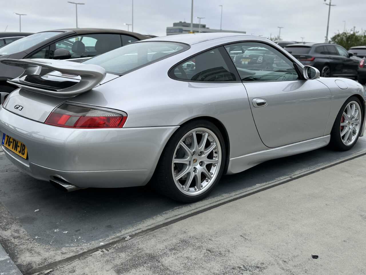Een zilveren Porsche 911 GT3 met achterspoiler staat op een bewolkte dag geparkeerd op een parkeerplaats. Op de achtergrond zijn andere auto's zichtbaar.