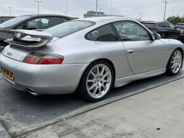 Een zilveren Porsche 911 GT3 met achterspoiler staat op een bewolkte dag geparkeerd op een parkeerplaats. Op de achtergrond zijn andere auto's zichtbaar.