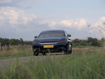 Een strakke zwarte BYD-auto met zichtbaar kenteken "S-314-NB" rijdt op een smalle weg omringd door groen gras en bomen onder een bewolkte hemel.