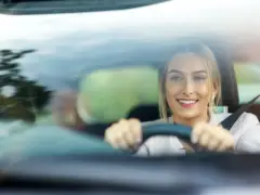 Een vrouw met blond haar glimlacht tijdens het rijden, gezien door de voorruit. Ze houdt het stuur met beide handen vast en draagt een veiligheidsgordel, en geniet duidelijk van haar tweede auto.