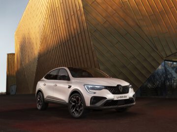 Een witte Arkana crossover SUV staat geparkeerd voor een eigentijds gouden gebouw met hoekige designelementen. De zonsondergang werpt een warme gloed op het toneel.