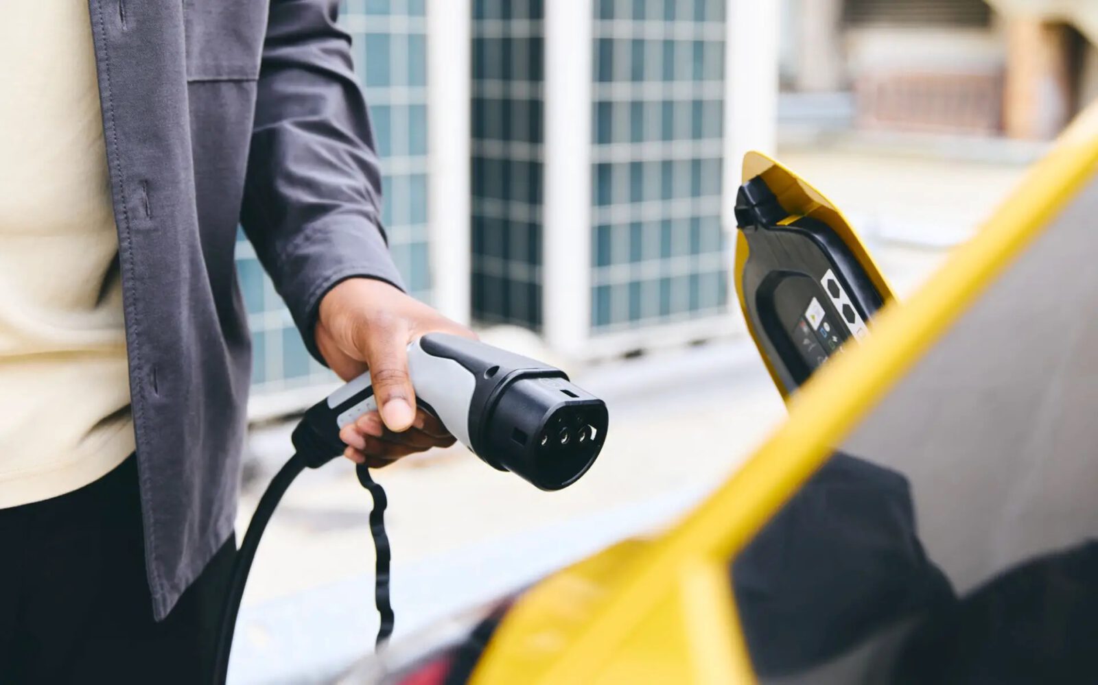 Een persoon houdt een oplaadstekker voor een elektrisch voertuig vast in de buurt van de open oplaadpoort van een gele auto, wat de overstap naar elektrisch rijden illustreert, ondanks zorgen dat dit duurder is dan traditionele brandstof.