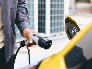 Een persoon houdt een oplaadstekker voor een elektrisch voertuig vast in de buurt van de open oplaadpoort van een gele auto, wat de overstap naar elektrisch rijden illustreert, ondanks zorgen dat dit duurder is dan traditionele brandstof.