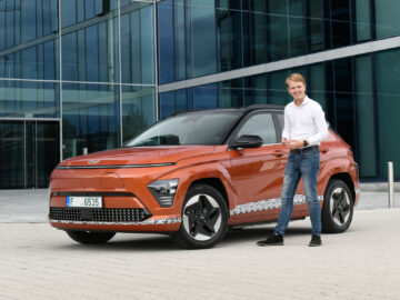 Een persoon staat naast een oranje Hyundai Kona Electric 2023, geparkeerd voor een modern glazen gebouw.