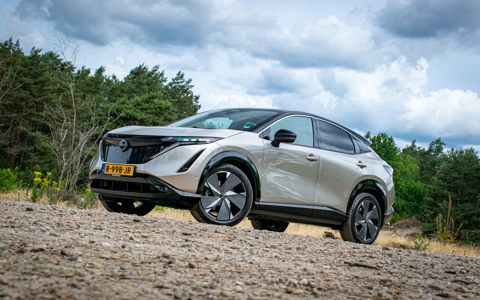 Een zilveren Nissan Ariya Engage SV met zwarte accenten staat geparkeerd op een grindpad, omgeven door een bos en een bewolkte lucht. Het voertuig pronkt trots met zijn Europese kenteken.
