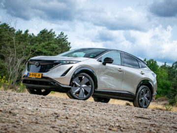 Een zilveren Nissan Ariya Engage SV met zwarte accenten staat geparkeerd op een grindpad, omgeven door een bos en een bewolkte lucht. Het voertuig pronkt trots met zijn Europese kenteken.