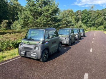 Vier kleine, identieke Opel Rocks Electric-auto's staan op een zonnige dag in een rij geparkeerd op een verharde weg omgeven door bomen en groen.