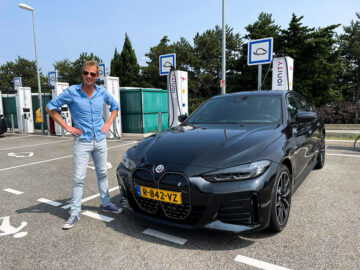 Op een zonnige zomervakantiedag staat iemand naast een zwarte BMW elektrische auto op een parkeerplaats. De auto heeft het kenteken "R-842-VZ." Op de achtergrond zijn verschillende borden en groen zichtbaar.