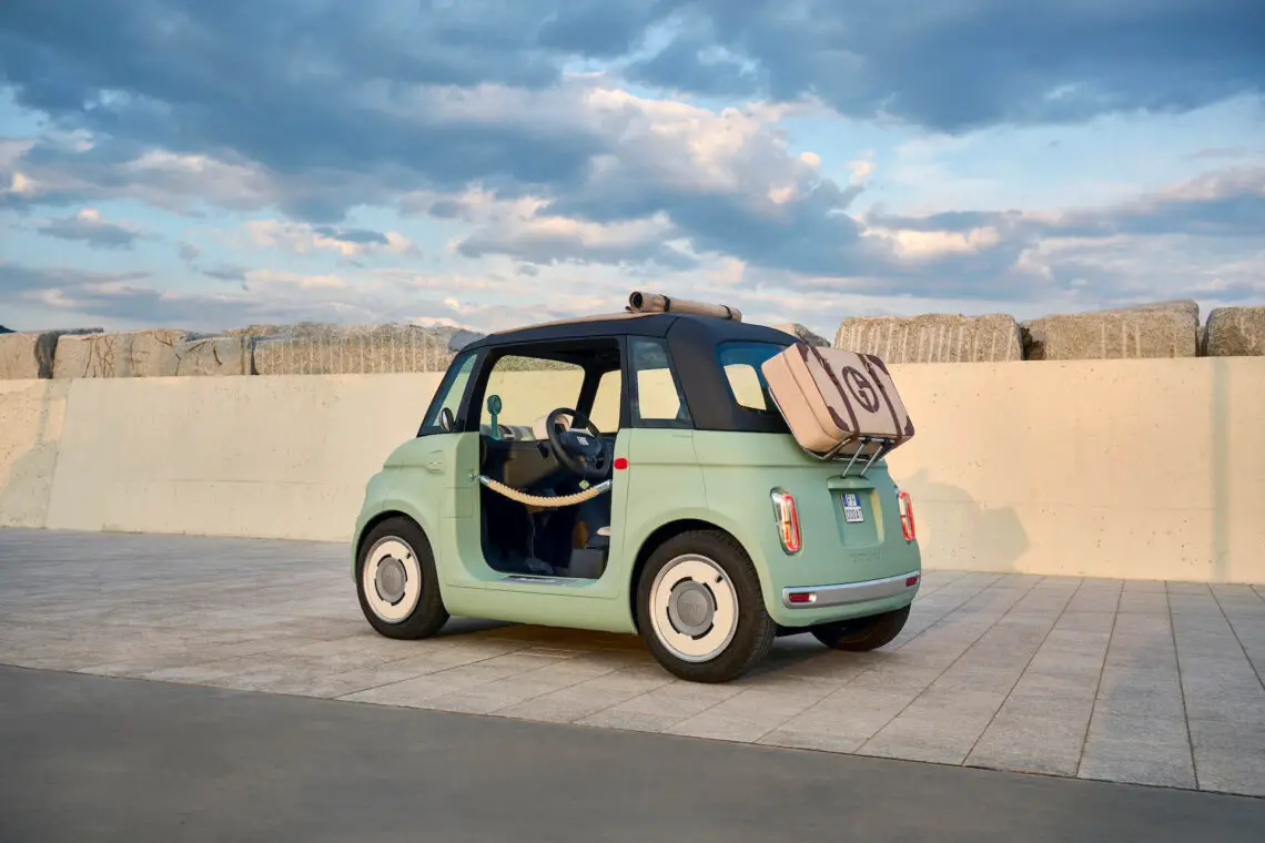 A FIAT Topolino compact electric car is parked on a paved lot near a concrete wall. The car has a light green exterior, a luggage compartment on the roof and an open driver's door that reveals the interior. Clouds fill the sky in the background.