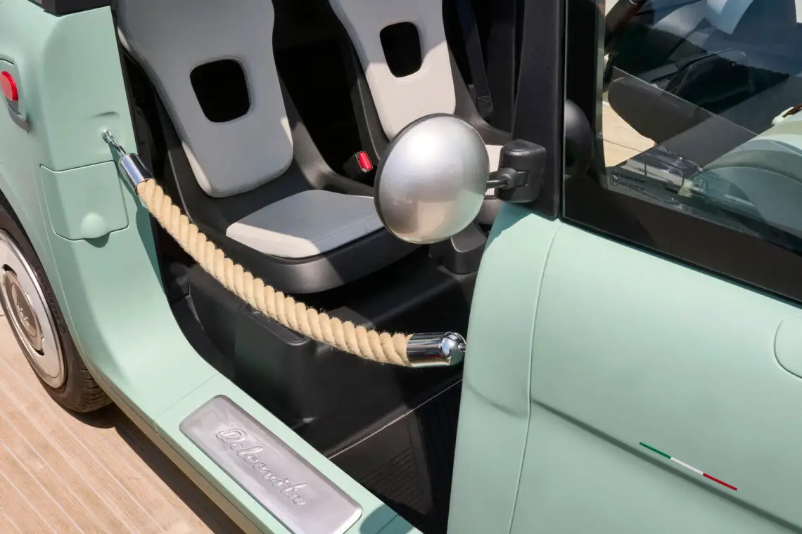 Close-up of the interior of a light green FIAT Topolino with two white seats, a rope barrier replacing the door and the Italian flag logo on the side.