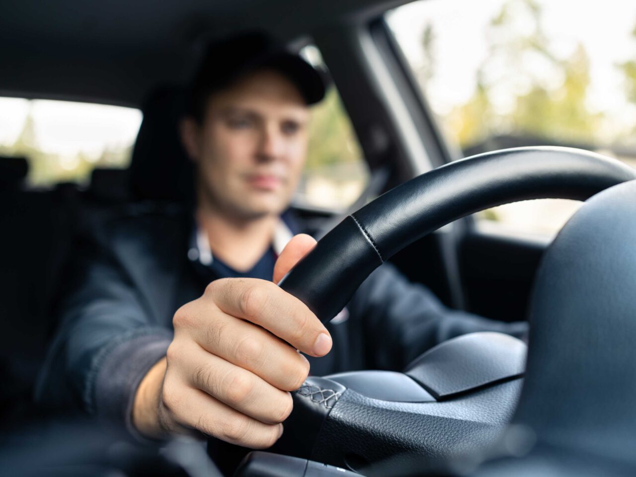 Een persoon bestuurt een auto, houdt het stuur met één hand vast en kijkt aandachtig vooruit. De bomen buiten vervagen naar de achtergrond en belichamen de betekenis van beweging en snelheid.