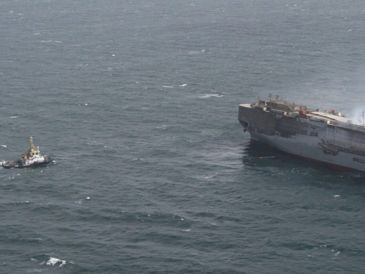 De sleepboot nadert de Fremantle Highway met rook die uit de boeg komt, gezien op open zee met schokkerige golven, wat duidt op een mogelijk brandscenario of een reddingsoperatie.