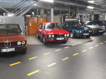 Binnen staat een opstelling van zes BMW-auto's uit verschillende tijdperken, waaronder de iconische BMW 5-serie. De voertuigen variëren van oudere klassieke modellen tot modernere versies en tonen de evolutie van BMW-design.