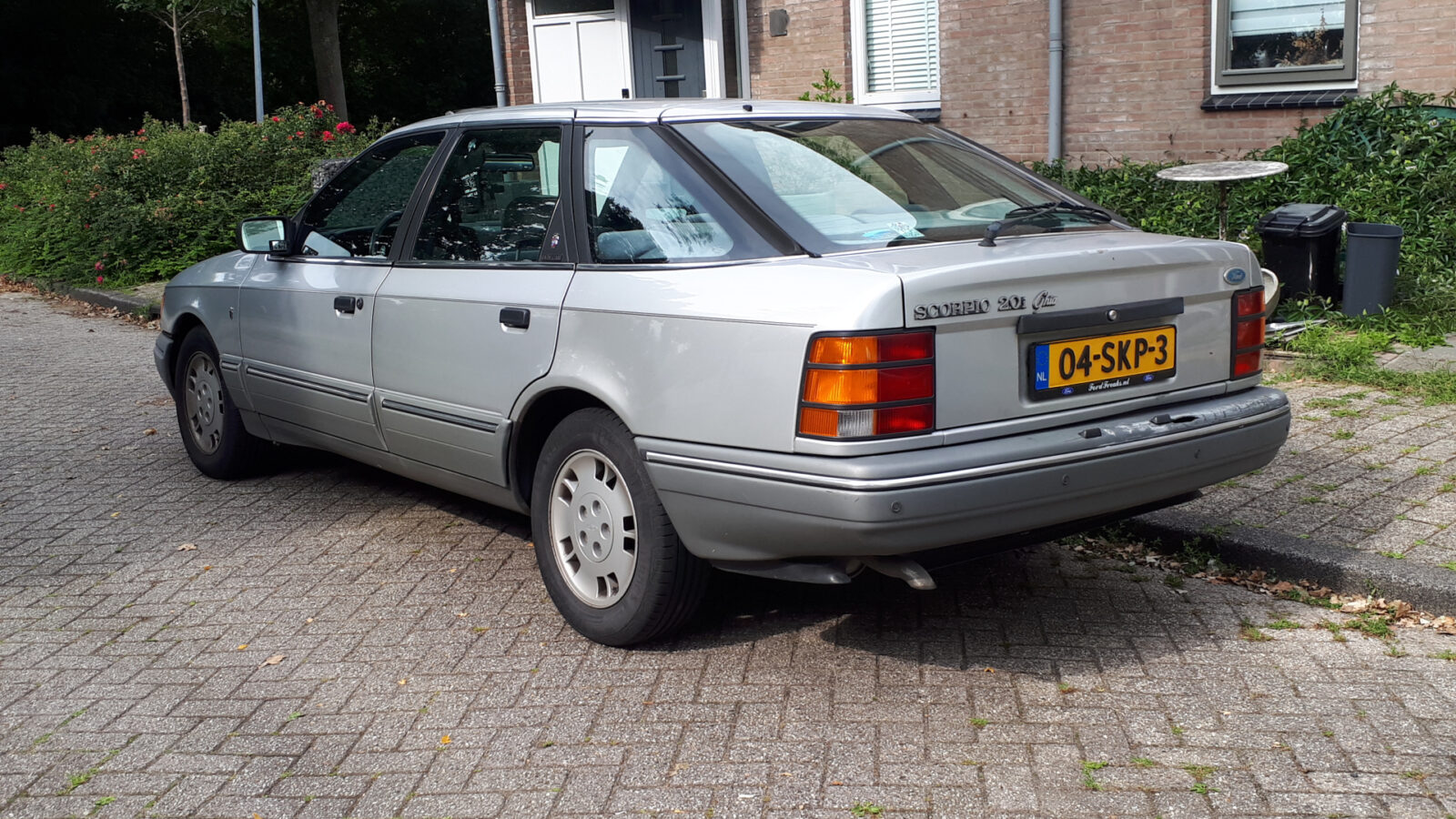 Een zilverkleurige Opel Vectra CD sedan uit de jaren 80 of 90 staat geparkeerd in een woonstraat, vlak naast een Ford Scorpio. De auto heeft een Nederlands kenteken met de vermelding 04-SK-P3. Struiken en een bakstenen gebouw bevinden zich op de achtergrond.