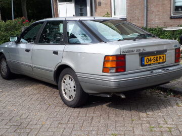Een zilverkleurige Opel Vectra CD sedan uit de jaren 80 of 90 staat geparkeerd in een woonstraat, vlak naast een Ford Scorpio. De auto heeft een Nederlands kenteken met de vermelding 04-SK-P3. Struiken en een bakstenen gebouw bevinden zich op de achtergrond.
