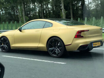 Een goudkleurige Polestar 1-sportwagen met een strak design en zwarte velgen rijdt over een weg omgeven door bomen en groen. Het kenteken is zichtbaar maar onleesbaar op de afbeelding.