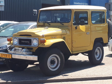 Een gele Toyota Land Cruiser staat geparkeerd in een zonnige straat naast andere voertuigen, waaronder een witte auto. Het robuuste ontwerp van de Land Cruiser valt op tegen de heldere achtergrond en benadrukt de ruige elegantie.