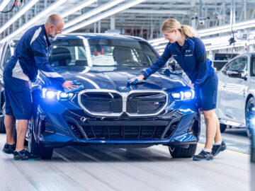 Twee arbeiders in blauwe uniformen voeren kwaliteitscontroles uit op een BMW 5 Serie-auto in een autoproductielijn.