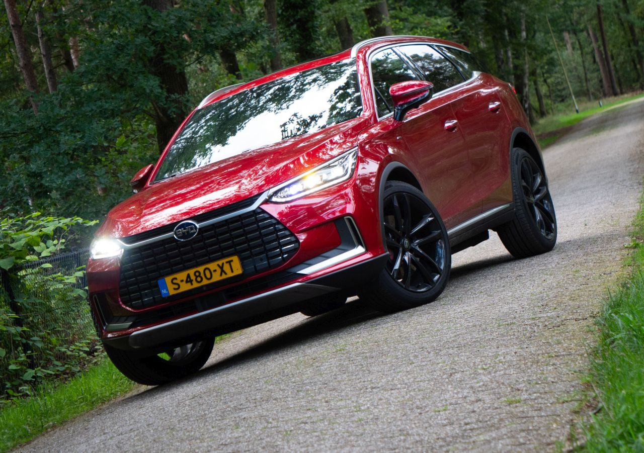 Een rode BYD Tang SUV met zwarte wielen staat geparkeerd op een smalle weg omgeven door bomen. Het voertuig heeft een Nederlands kenteken, S-480-XT.