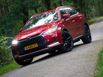 Een rode BYD Tang SUV met zwarte wielen staat geparkeerd op een smalle weg omgeven door bomen. Het voertuig heeft een Nederlands kenteken, S-480-XT.