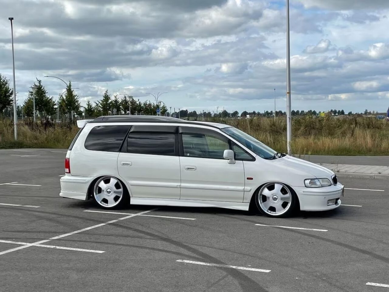 Een witte Honda Odyssey-minibus met aangepaste lage vering en grote zilveren velgen staat geparkeerd op een lege parkeerplaats met een bewolkte lucht en een grasrijke omgeving op de achtergrond.