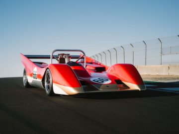 Een vintage rood-witte Lotus Type 66-raceauto met nummer 66, met een grote achtervleugel, geparkeerd op een racecircuit tegen een helderblauwe lucht.