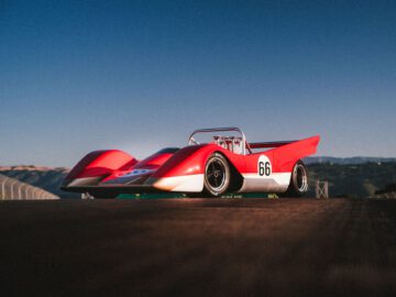 Een rood-witte vintage Lotus Type 66-raceauto met het nummer 66 op de zijkant staat geparkeerd op een lege, zonovergoten baan onder een helderblauwe hemel.