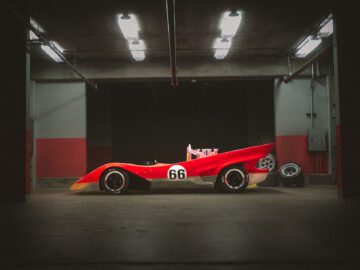 Een rood-witte raceauto, de Lotus Type 66 met het nummer 66 erop geschilderd, staat geparkeerd in een slecht verlichte garage met enkele banden op de vloer.